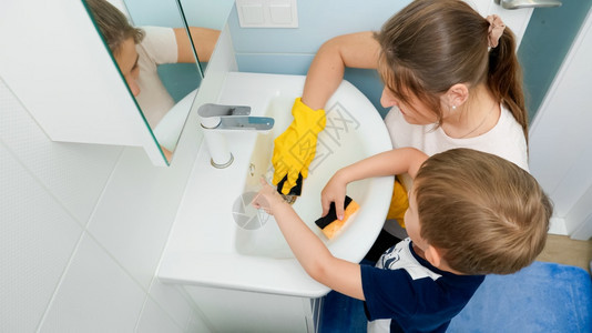 快乐的男孩母亲戴着橡皮手套在海绵上涂洗涤剂和清洁洗浴室水池小男孩帮助父母做家务快乐的男孩在海绵上涂洗涤剂和浴室水池图片