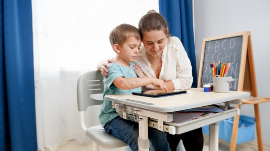年轻母亲拥抱在家中做功课和平板电脑上学习的小儿子Child使用教育和学习的工具图片