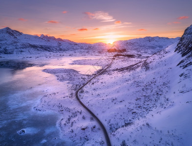 风向道路雪覆盖山冬季日落时橙色天空的中景象海冰冷岸雪岩公路洛福滕岛挪威冷冻海岸背景图片