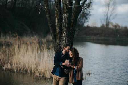 有两个天主教情人站在湖边的树旁年轻夫妇在秋天户外拥抱一个有胡子的男人和卷毛女在爱中情节爱的概念两个天主教情人站在湖边的树年轻夫妇图片
