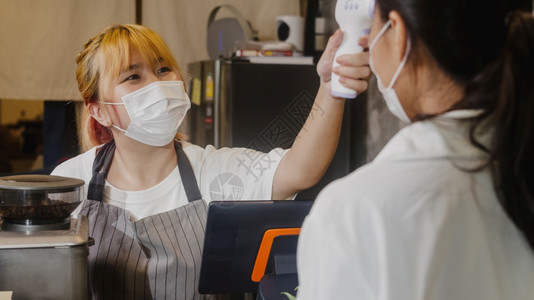 年轻亚洲女餐厅员工在进入前额戴保护面罩,使用红外温度计检查器或对客户和’;进入前额。图片
