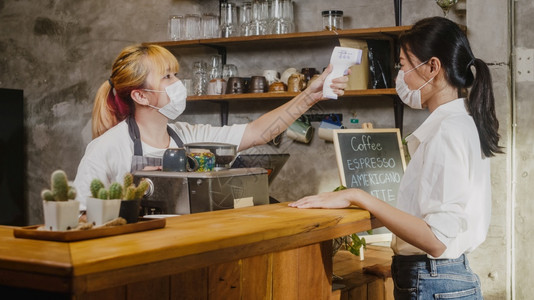 年轻亚洲女餐厅员工在进入前额戴保护面罩,使用红外温度计检查器或对客户和’;进入前额。图片