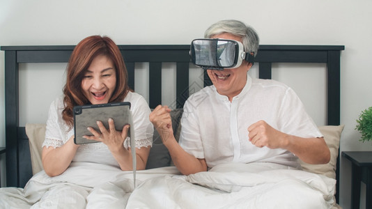 亚裔老人夫妇在家里使用平板电脑亚裔老年人祖父母在早晨的观念下躺在卧室的床上时一起玩游戏感到快乐和VR玩游戏图片