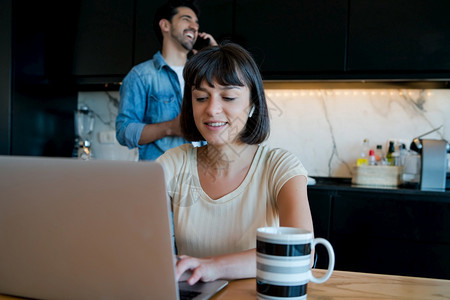 年轻女在家里用笔记本电脑工作的肖像而男则在背景上通过电话交谈家庭办公室的概念新正常生活方式图片