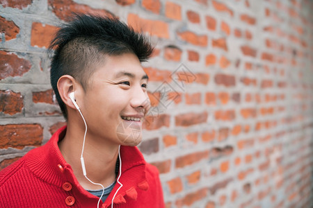 在户外用耳机和砖墙对着听音乐的亚洲男孩肖像城市概念图片