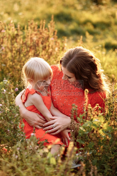 美丽的年轻人和女儿坐在草地上与日落时的夏绿田相对母亲日妇女微笑的年轻美人和女儿坐在草地上与日落时的夏绿地对立图片