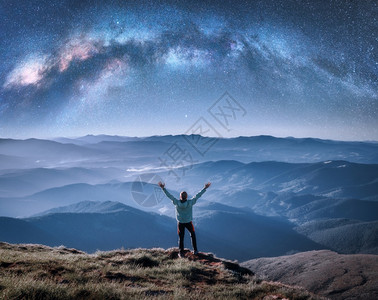 在山顶上欢乐的男子在山顶上横跨脉的银河在夜晚低云的中蓝天空的风景与星银河拱门小伙子雾中的山丘空间和星系空与恒的天图片