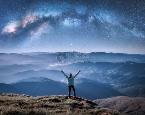 在山顶上欢乐的男子在山顶上横跨脉的银河在夜晚低云的中蓝天空的风景与星银河拱门小伙子雾中的山丘空间和星系空与恒的天图片