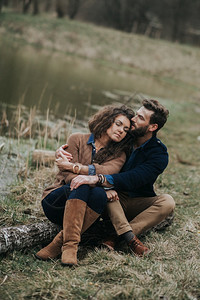 快乐的情侣坐在湖岸边年轻夫妇在秋天的户外拥抱一个有胡子的男人和卷毛女在爱中情节爱与家庭的概念快乐情侣坐在湖岸边年轻夫妇在秋天的户图片