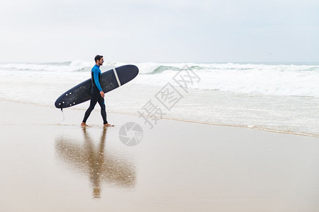 年轻男冲浪者穿着泳衣手臂下握着冲浪板早上后在海滩散步图片