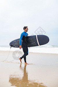 年轻男冲浪者穿着泳衣手臂下握着冲浪板早上后在海滩散步图片