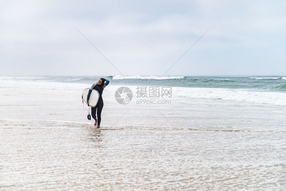 年轻女冲浪者穿着泳衣手臂下握着冲浪板早上后在海滩散步图片