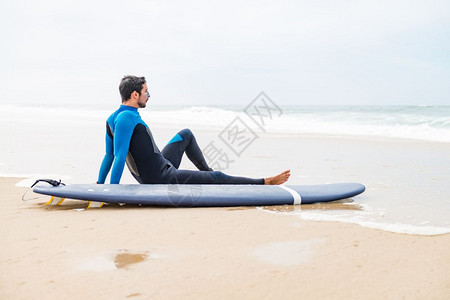 年轻男冲浪者穿着泳衣早上冲浪课后坐在海滩上的冲浪板旁背景图片
