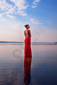 穿着红裙子的迷人女孩站在水中美丽日出的背景下美丽时尚户外图片