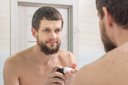 一个未剃毛的人用须泡沫在他的手掌上看着在浴室的镜子图片