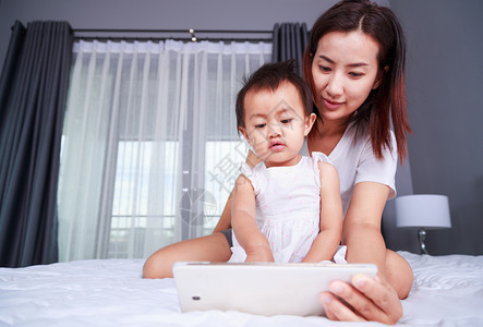母亲和婴儿正在家里的床上玩耍和阅读平板电脑图片