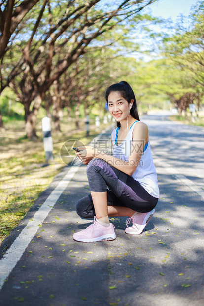 坐在公园路边的年轻运动妇女坐在公园的路上并用有耳机的智能手监听音乐图片