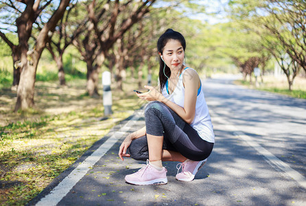 坐在公园路边的年轻运动妇女坐在公园的路上并用有耳机的智能手监听音乐图片