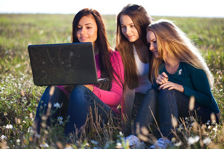 三位美丽的年轻女士手持笔记本电脑坐在天空背景的田地上图片
