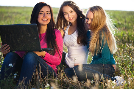 三位美丽的年轻女士手持笔记本电脑坐在天空背景的田地上图片