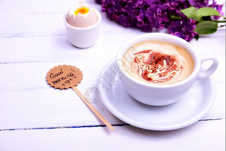 一杯卡布奇诺咖啡白色木质背景上有一个茶托后面是一束紫丁香图片