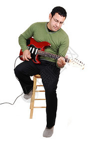 一个中年男子坐在工作室里弹吉他穿着衣服裤子和绿色毛衣白背景图片