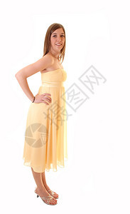 一个漂亮的年轻女人穿着长黄色的裙子穿着高跟鞋站在工作室里微笑在镜头站白色背景上图片