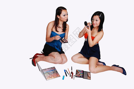 两位亚裔女坐在摄影棚的地板上帮助对方为下一张图片拍摄化妆以浅灰色背景拍摄图片