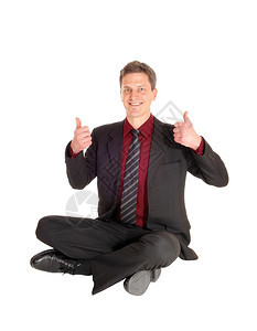 一个穿着西装的商人坐在地板上两只拇指都举起来穿着瑜伽姿势孤立在白色背景上图片
