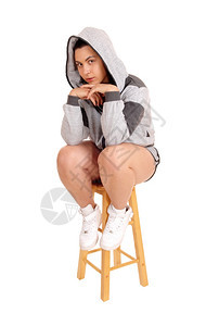 一个穿着灰色连帽衫和短裤的可爱年轻女子坐在椅上手放下巴因为白色背景而孤立无援图片