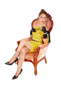 一位可爱的年轻女子穿着黄色礼服坐在粉红色的扶手椅上拿着长的烟筒与白种背景隔绝图片