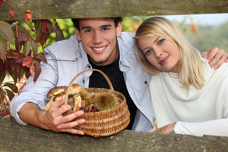 秋天在木制屏障后面的一对年轻夫妇男子拿着一篮里面有蘑菇和栗子图片