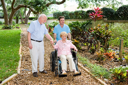 坐在轮椅上的年长妇女正由一名修士及其丈夫步行穿过疗养院花园图片