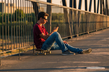 年轻人坐在滑板上河边的行人桥上发短信图片