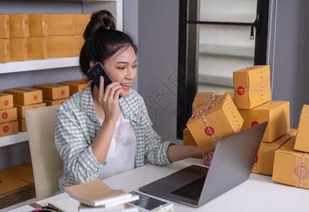 年轻妇女在智能手机上讲话使用电脑笔记本在家庭办公室网上销售产品图片