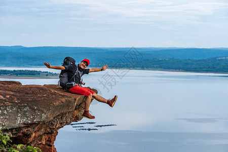 欢乐的徒步男子坐着和手势举起臂在悬崖边缘岩石山顶图片