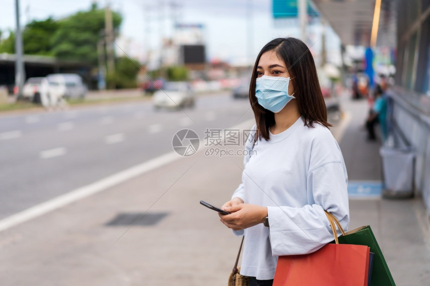 年轻女手持智能机和购物袋在市街公共汽车站等并戴面罩保护传播新正常概念的冠状新冠流行病图片