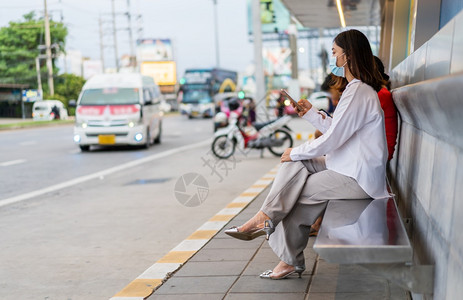 持有智能手机在市街公交站等车的年轻女青妇并戴面罩保护传播新正常概念的冠状新冠流行病图片