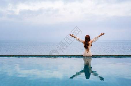 坐在游泳池边缘双臂举起的年轻女子仰望着大海图片