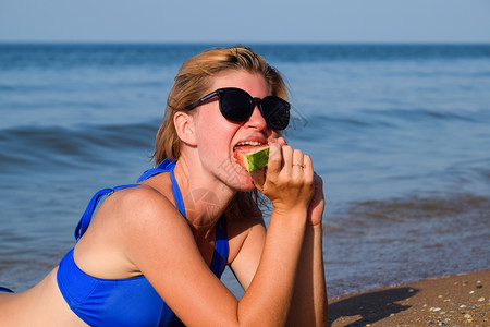身着太阳镜的金发女郎躺在海浪附近的滩上吃西瓜一只多汁的西瓜在一个女人的手里一个身着太阳镜的金发女郎躺在海浪附近的滩上一只多汁的西图片