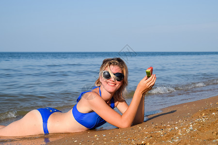 身着太阳镜的金发女郎躺在海浪附近的滩上吃西瓜一只多汁的西瓜在一个女人的手里一个身着太阳镜的金发女郎躺在海浪附近的滩上一只多汁的西图片