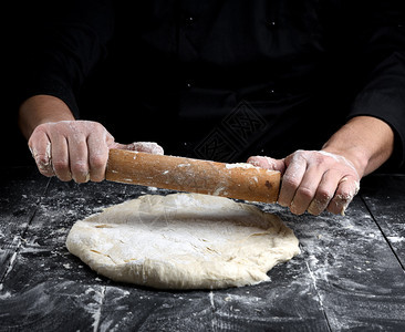 黑外衣厨师在桌上用木滚动针做圆披萨的面团图片