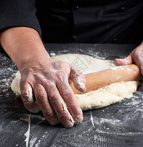 黑外衣厨师在桌上用木滚动针做圆披萨的面团图片