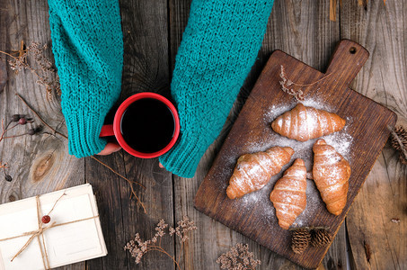 身着绿色编织毛衣的妇女手握着一个红瓷杯黑咖啡旁边是涂满糖粉灰木背景的烤羊角面包图片