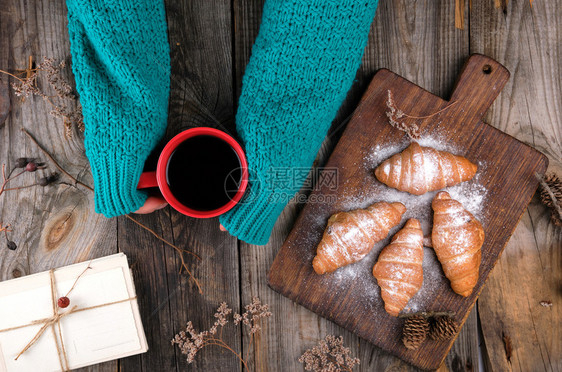 身着绿色编织毛衣的妇女手握着一个红瓷杯黑咖啡旁边是涂满糖粉灰木背景的烤羊角面包图片