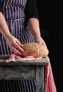 身穿蓝条纹围裙的妇女手握着用红麦面包和向日葵籽木制桌图片