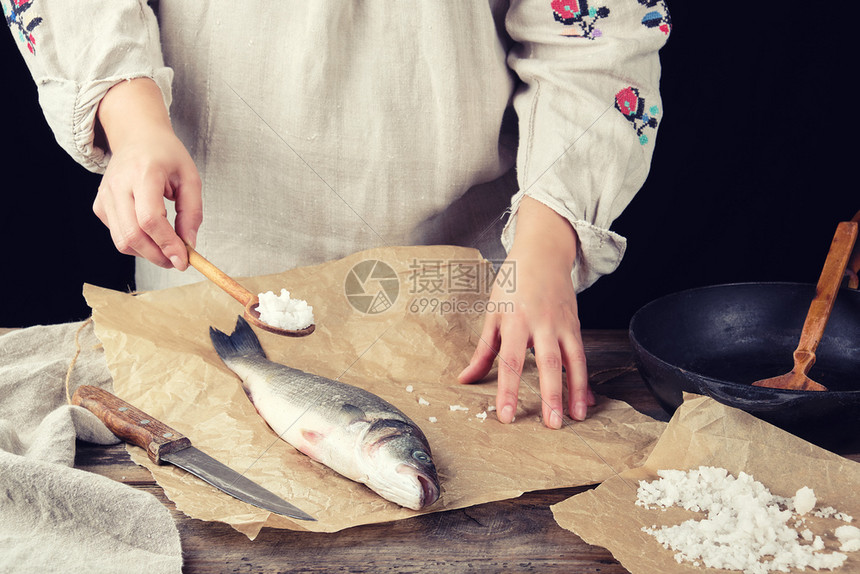 新鲜的整条海鲈鱼躺在棕色的纸上穿着灰色亚麻长裙的女孩拿着一勺盐黑色的背景图片