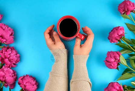 红奶油杯黑咖啡在女手中和一束红色的花朵绿叶子在蓝背景顶层视野图片