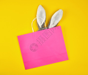 黄色背景的白手柄长角粉红纸购物袋兔子耳朵从里伸出来平躺着图片