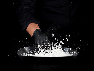 身穿黑色制服的主厨拿着一个有白盐的锅子吐出有选择焦点模糊调味粒子背景图片